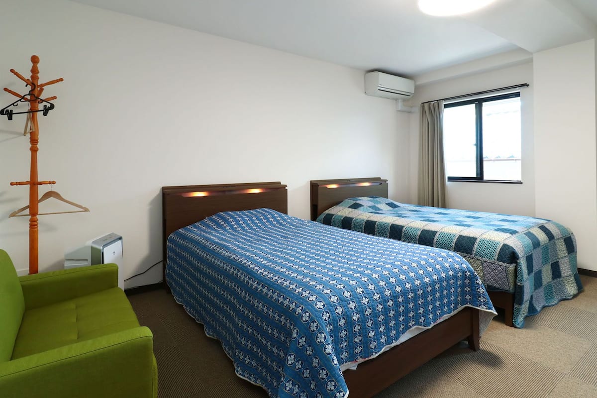 Habitación de hotel con dos camas, una silla verde, un equipo de aire acondicionado y una ventana con cortinas.
