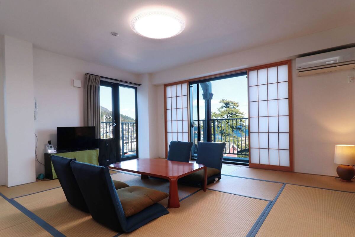 Habitación de hotel con suelo de tatami, una mesa baja, dos sillas, un televisor y una puerta corredera a un balcón.