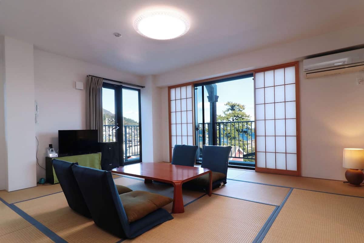 A hotel room with tatami flooring, a low table, two chairs, a TV, and a sliding door to a balcony.