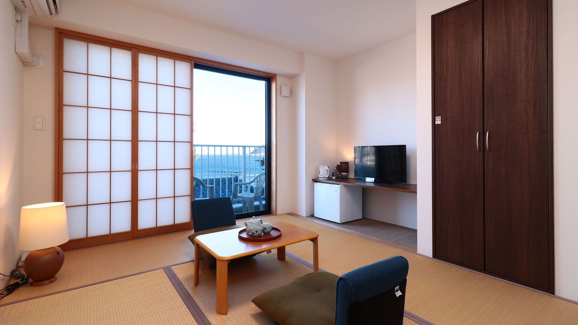 Una habitación con suelo de tatami, un panel corredero shoji, televisor, refrigerador pequeño y vista al mar.