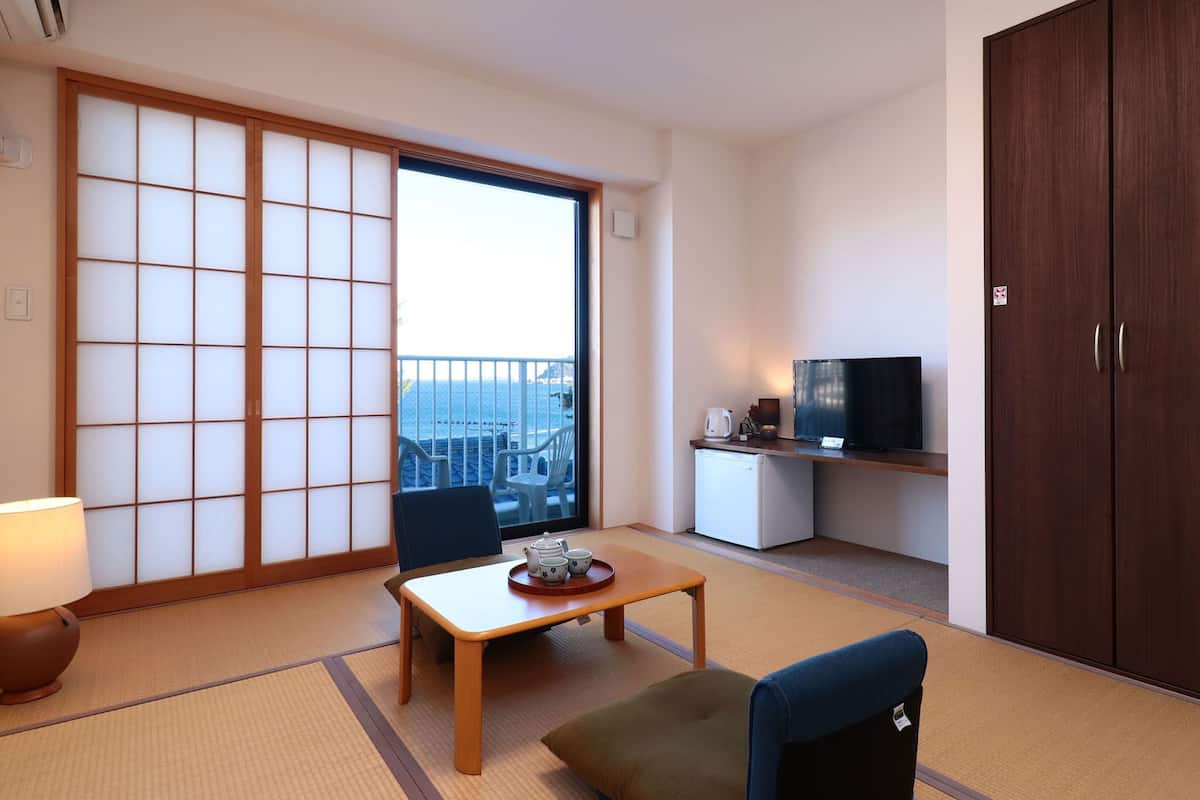 Una habitación con suelo de tatami, un panel corredero shoji, televisor, refrigerador pequeño y vista al mar.