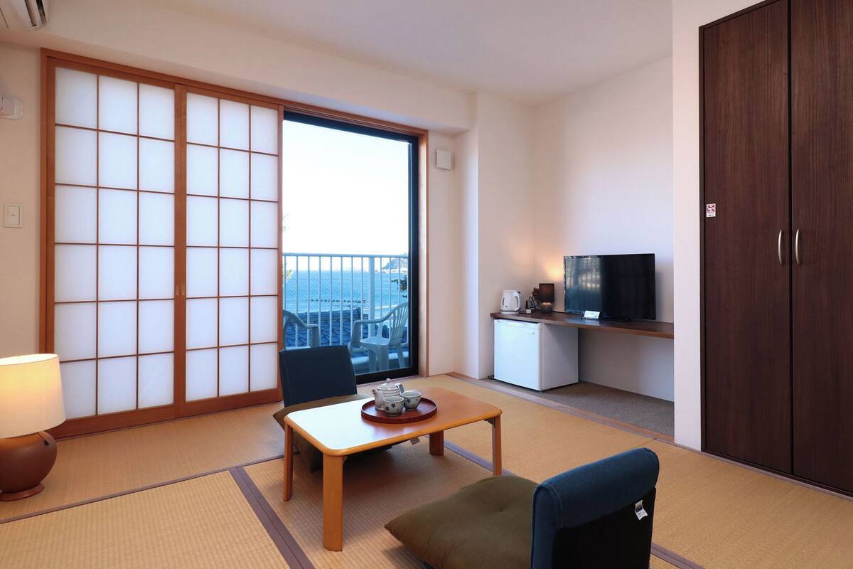 Una habitación con suelo de tatami, una puerta corredera estilo shoji, televisor, refrigerador pequeño y vista al mar.