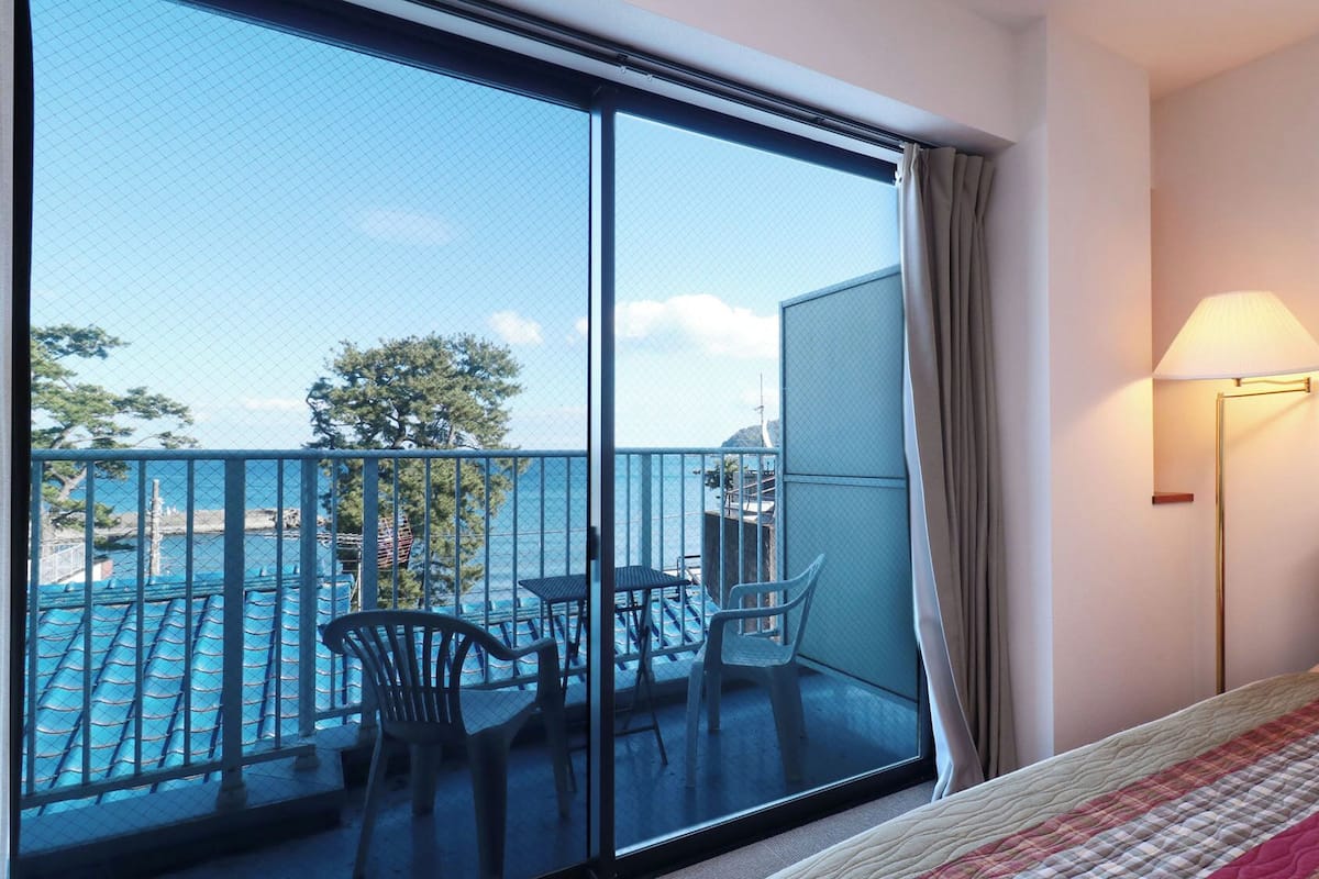 Habitación de hotel con un balcón grande, una cama, una mesa con sillas y una lámpara.