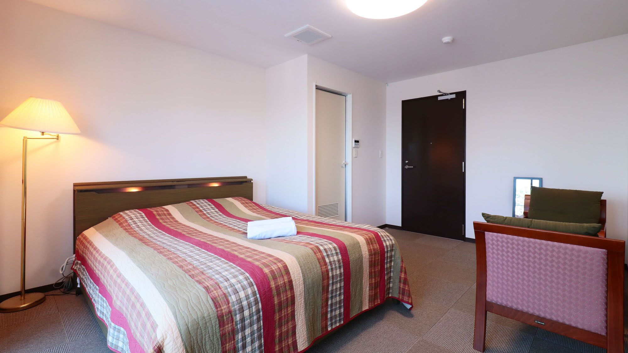 Una habitación de hotel con cama, lámpara de noche, una silla y una puerta.