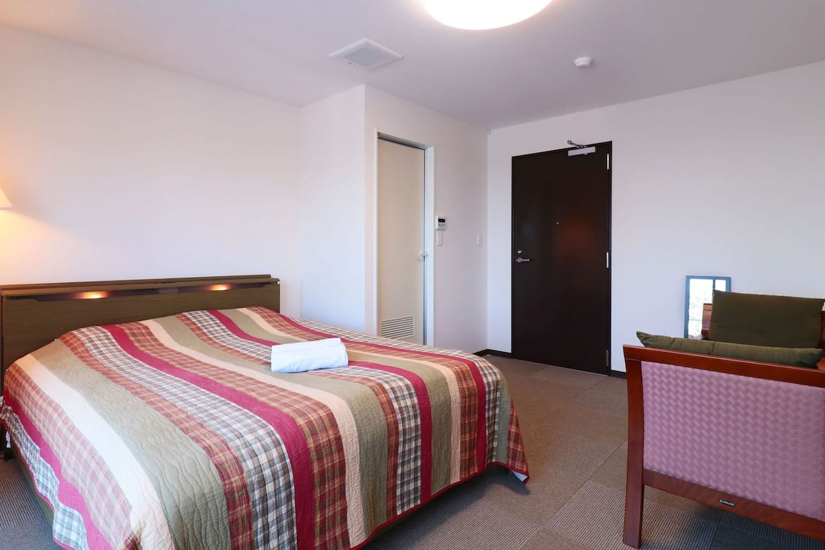 Una habitación de hotel con cama, lámpara de noche, una silla y una puerta.