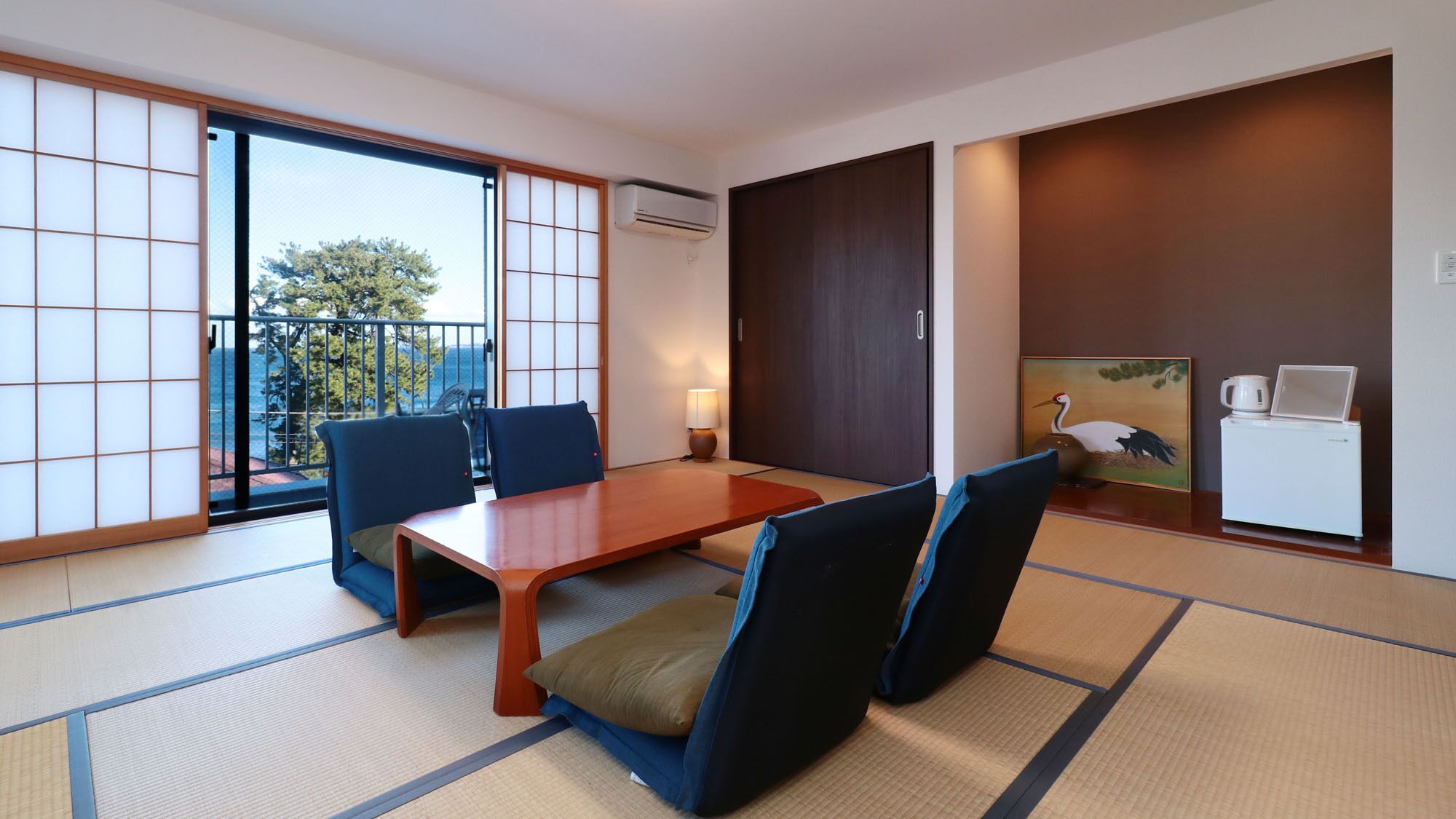 Una habitación japonesa tradicional con suelo de tatami, puertas correderas, una mesa con sillas, una lámpara, un cuadro y un refrigerador.