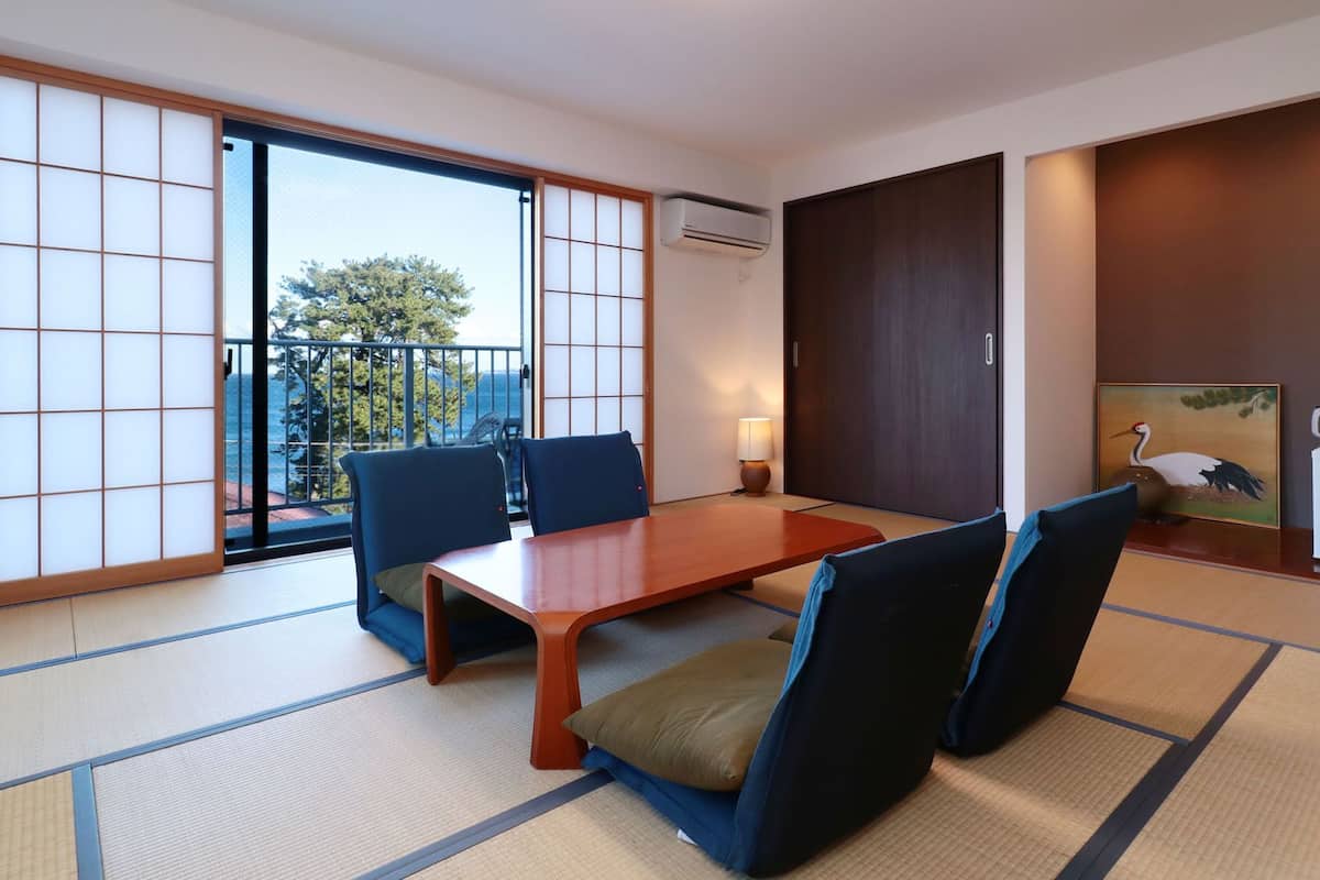 Una habitación japonesa tradicional con suelo de tatami, puertas correderas, una mesa con sillas, una lámpara, un cuadro y un refrigerador.