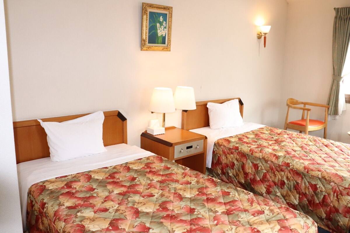A hotel room with two beds, a nightstand with lamps, a chair, and a wall-mounted picture.