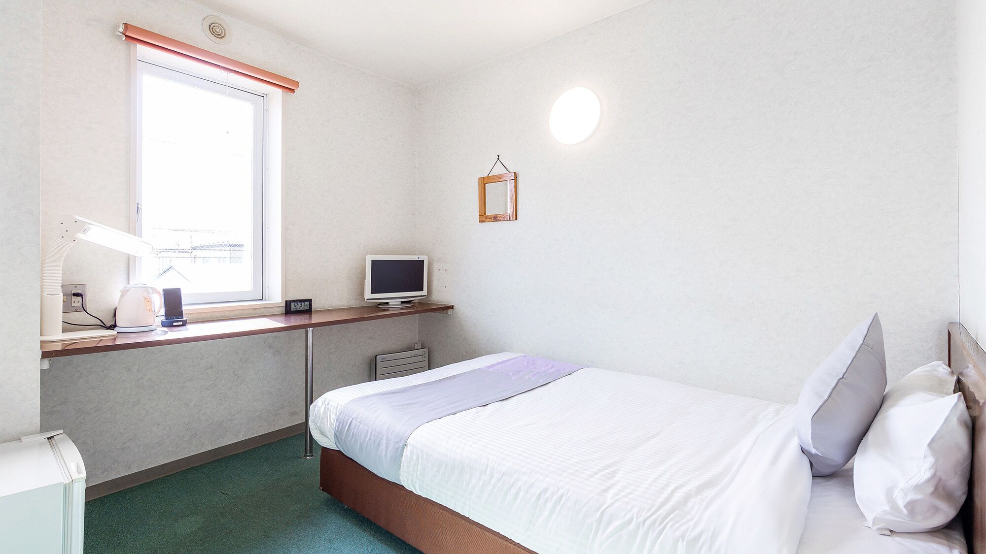 Een slaapkamer met een bed, een bureau met een computer, een raam en een wandlamp.