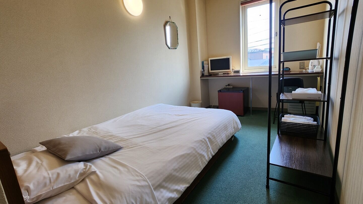 Een enkel bed met een kussen, een bureau met een computer, een stapelbed en een raam met jaloezieën.