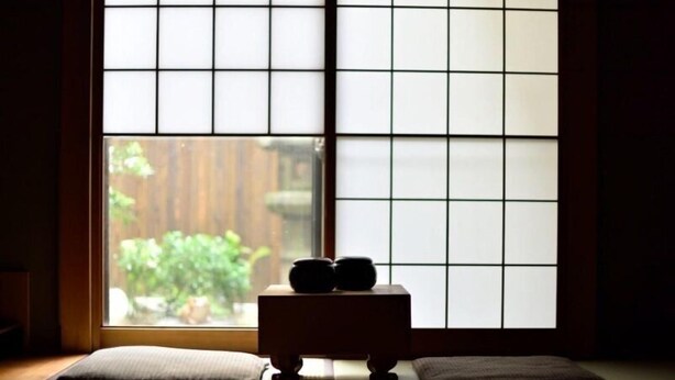 Une pièce avec un sol recouvert de tatamis, une cloison coulissante de style shoji, une petite table en bois sur laquelle reposent deux objets noirs, et un coussin.