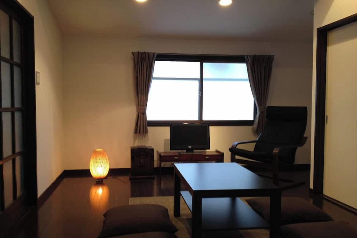 A room with a bed, a TV, a chair, a table, and a window with curtains.