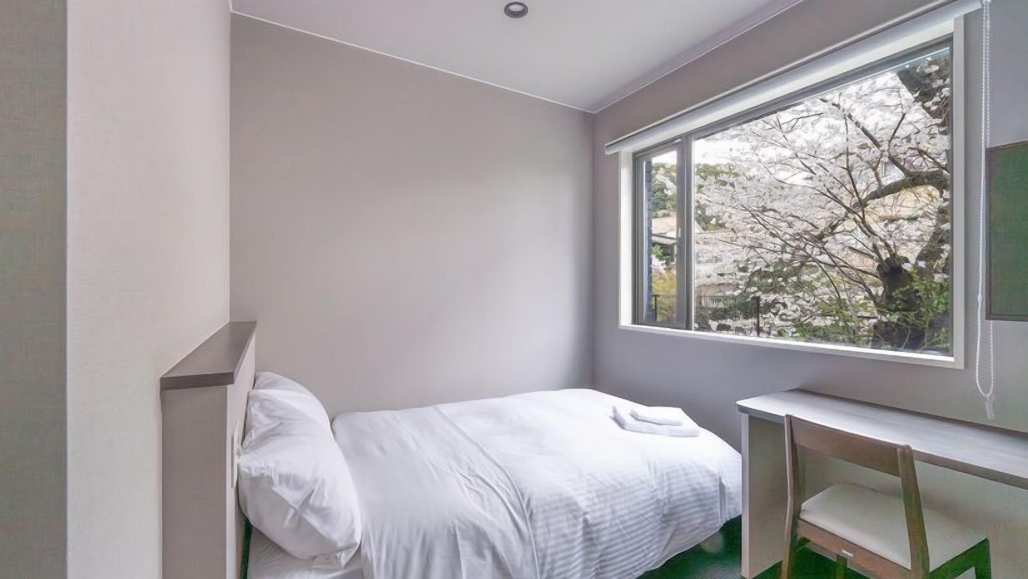 A small, modern bedroom with a single bed, a desk, and a large window offering a view of trees.