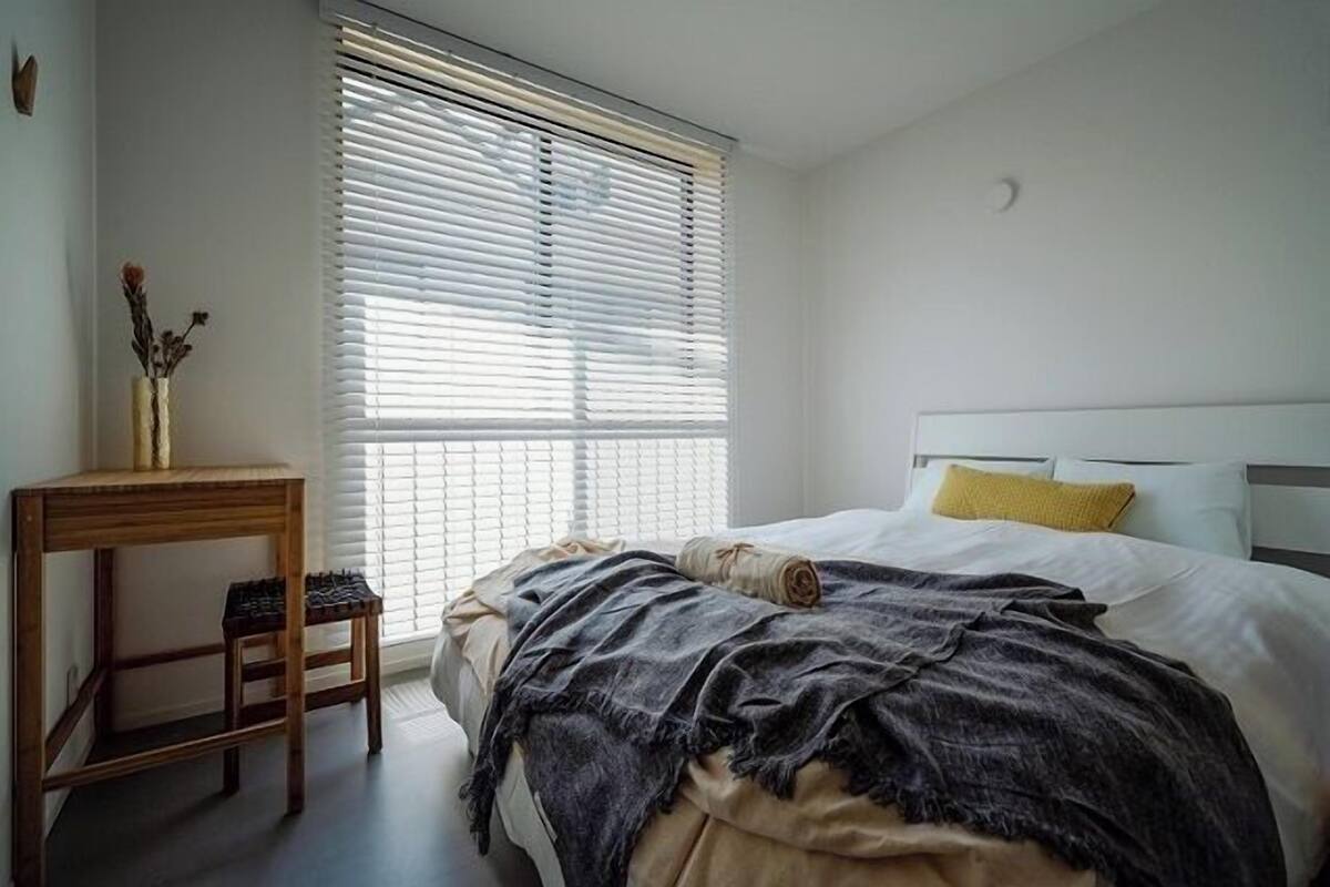 Un dormitorio con cama, mesita de noche, ventana con persianas y un taburete pequeño.