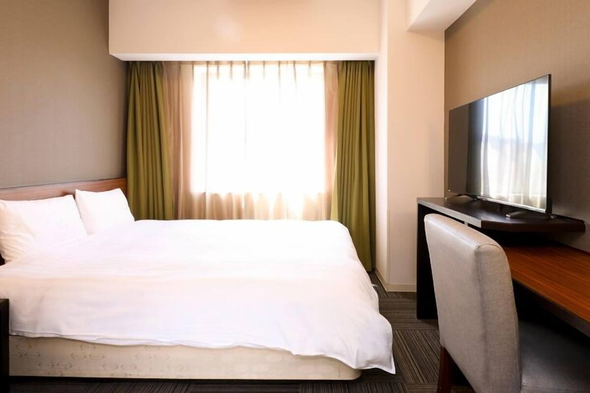 A hotel room with a bed, a desk with a television, and a window with curtains.