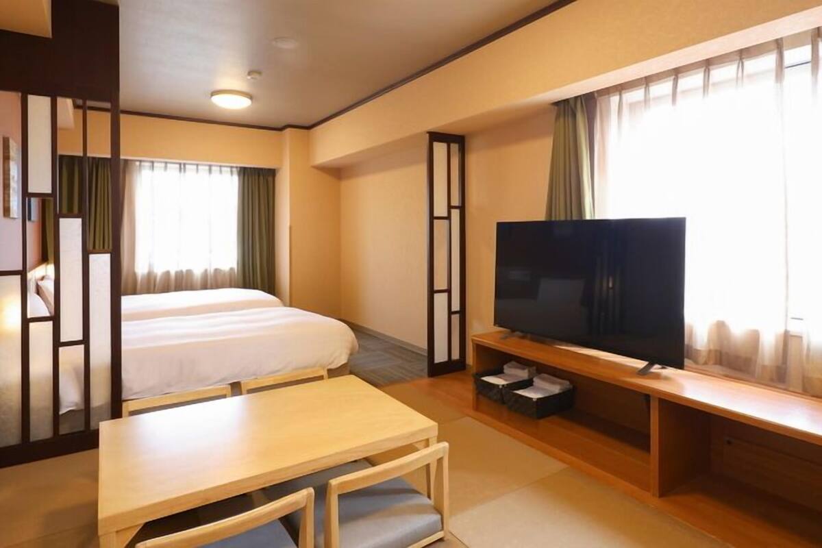 A hotel room with a bed, a TV, a desk, and a window with curtains.
