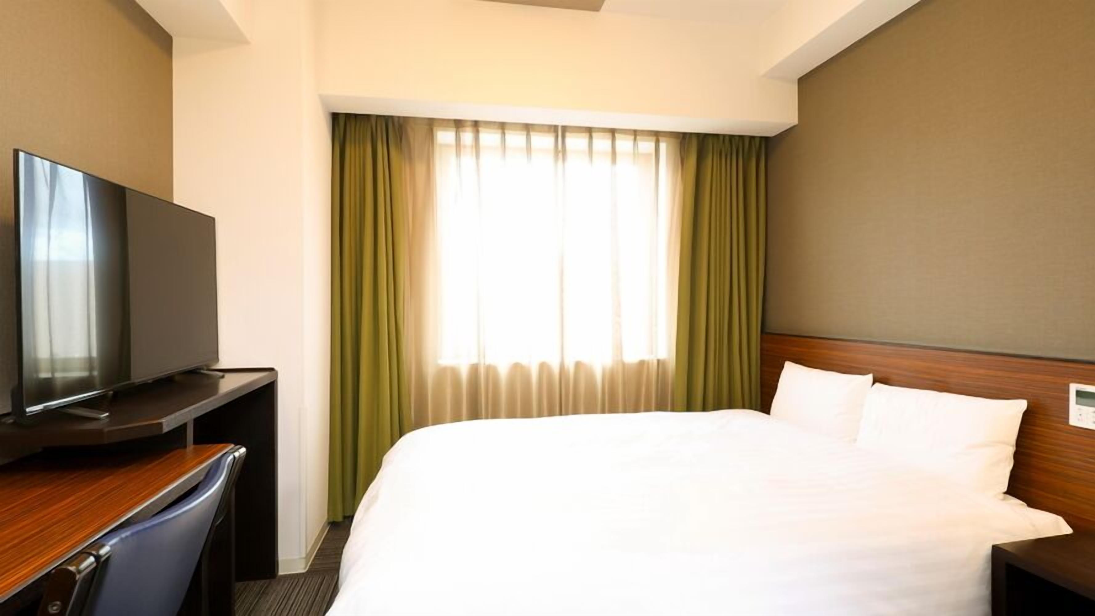 A hotel room with a bed, a TV on a desk, and a window with curtains.