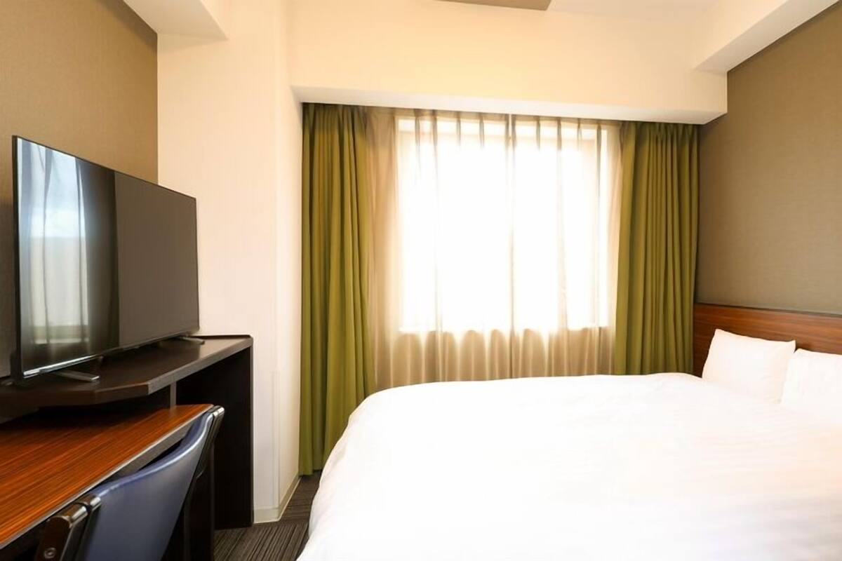A hotel room with a bed, a TV on a desk, and a window with curtains.