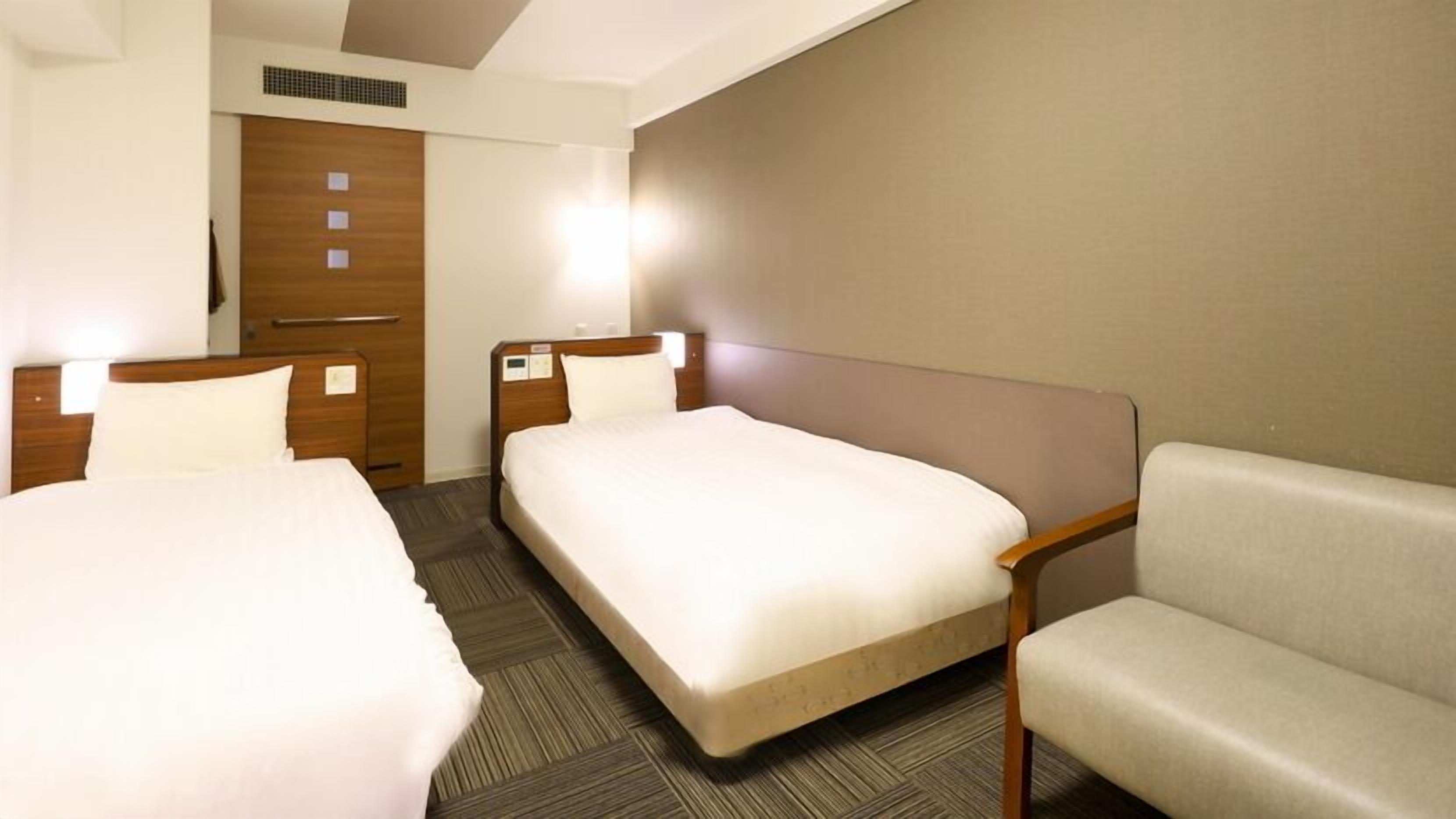 A hotel room with two beds, a sofa, a wooden wardrobe, and a wall-mounted heater.