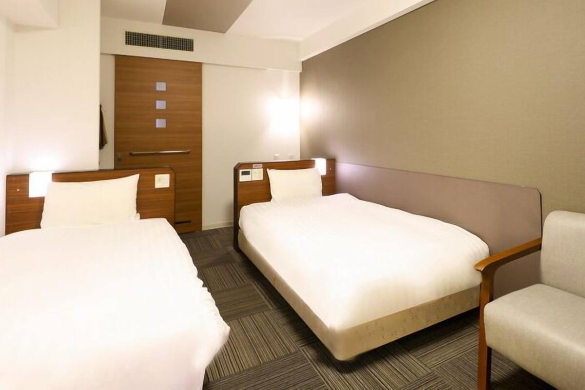 A hotel room with two beds, a sofa, a wooden wardrobe, and a wall-mounted heater.
