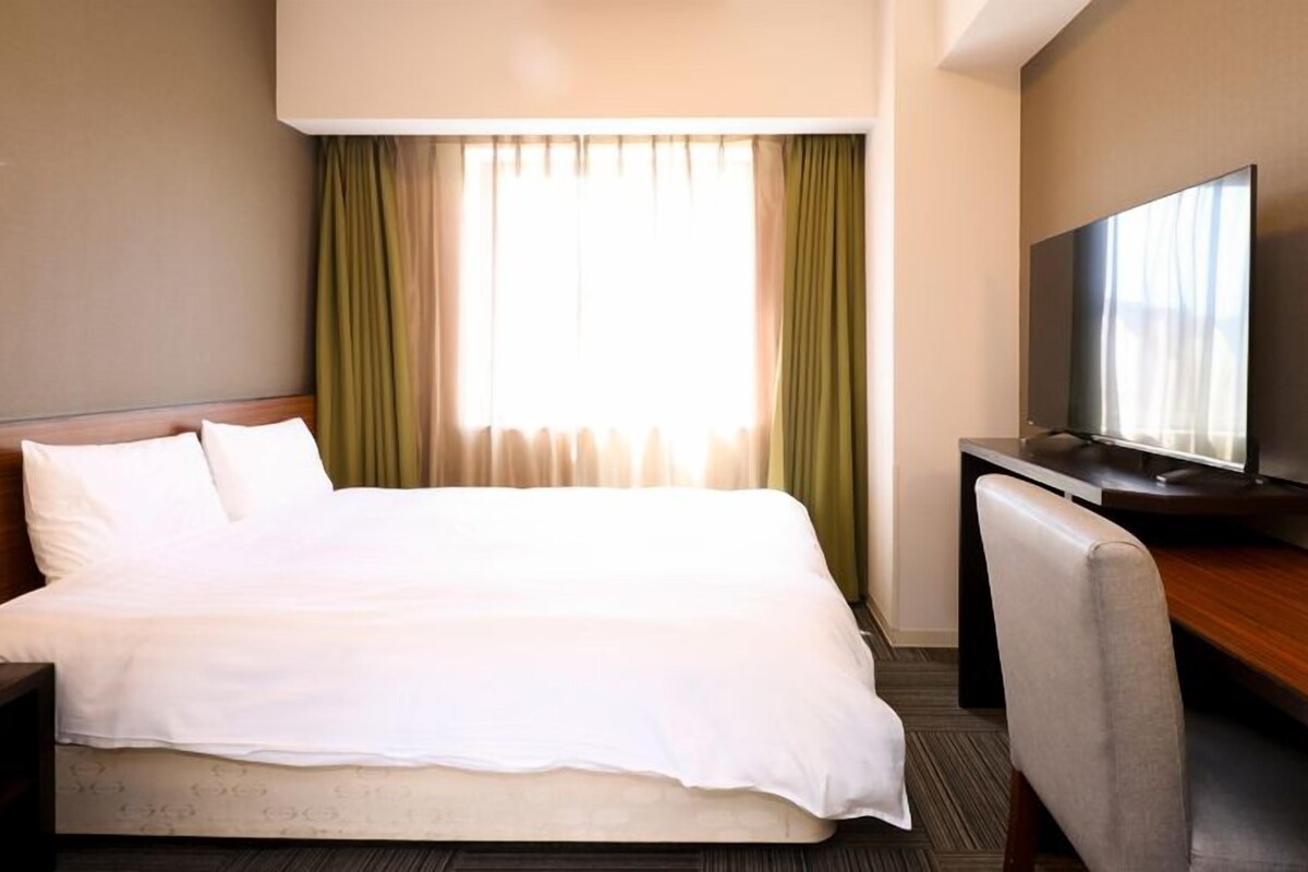 A hotel room with a bed, a desk with a television, and a window with curtains.