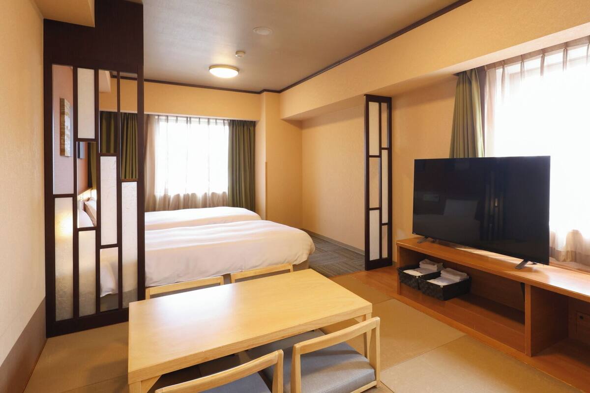 A hotel room with a bed, a TV, a desk, and a window with curtains.