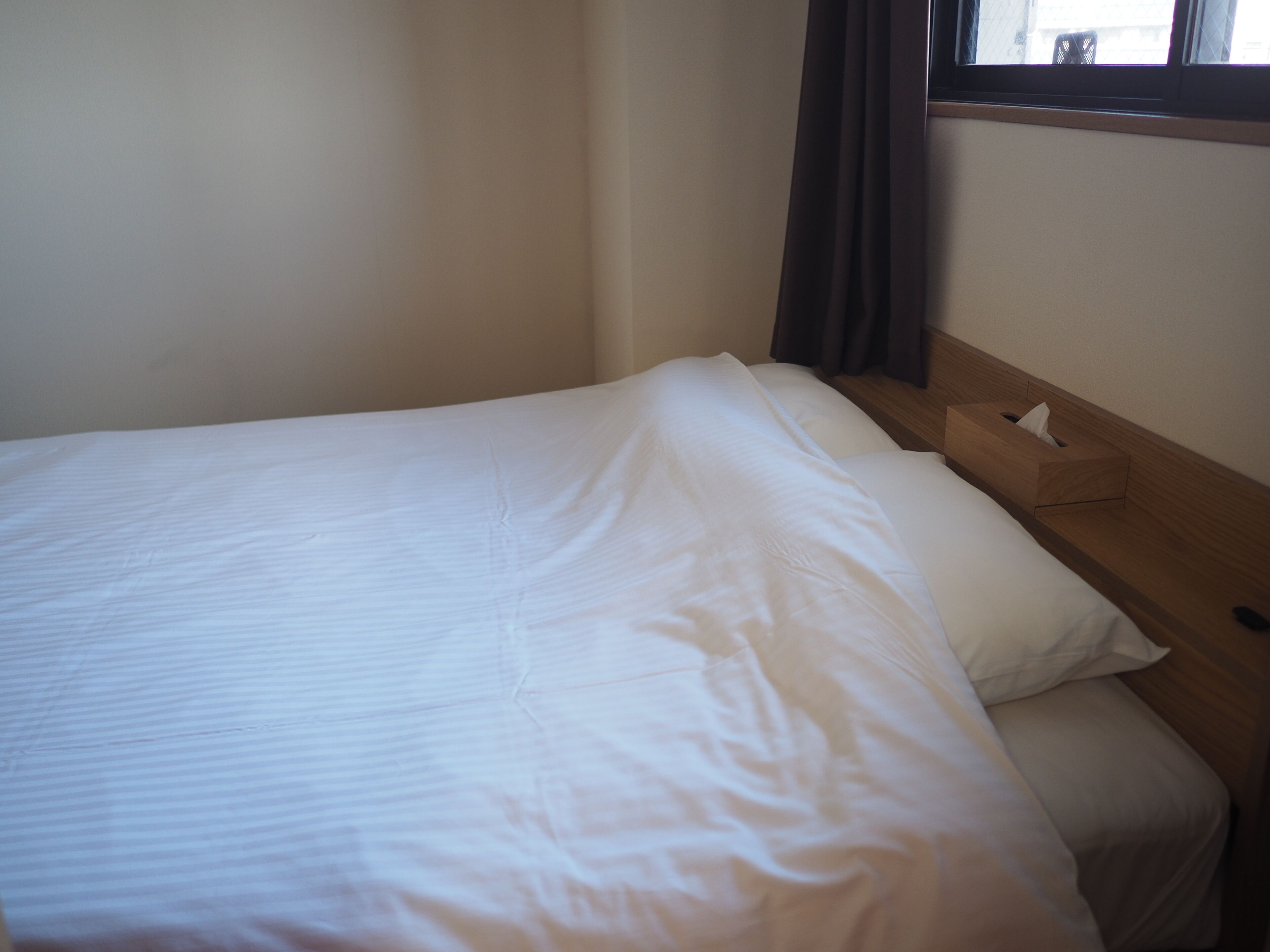 A single bed with a wooden headboard, a bedside table with a tissue box, and a window with curtains.
