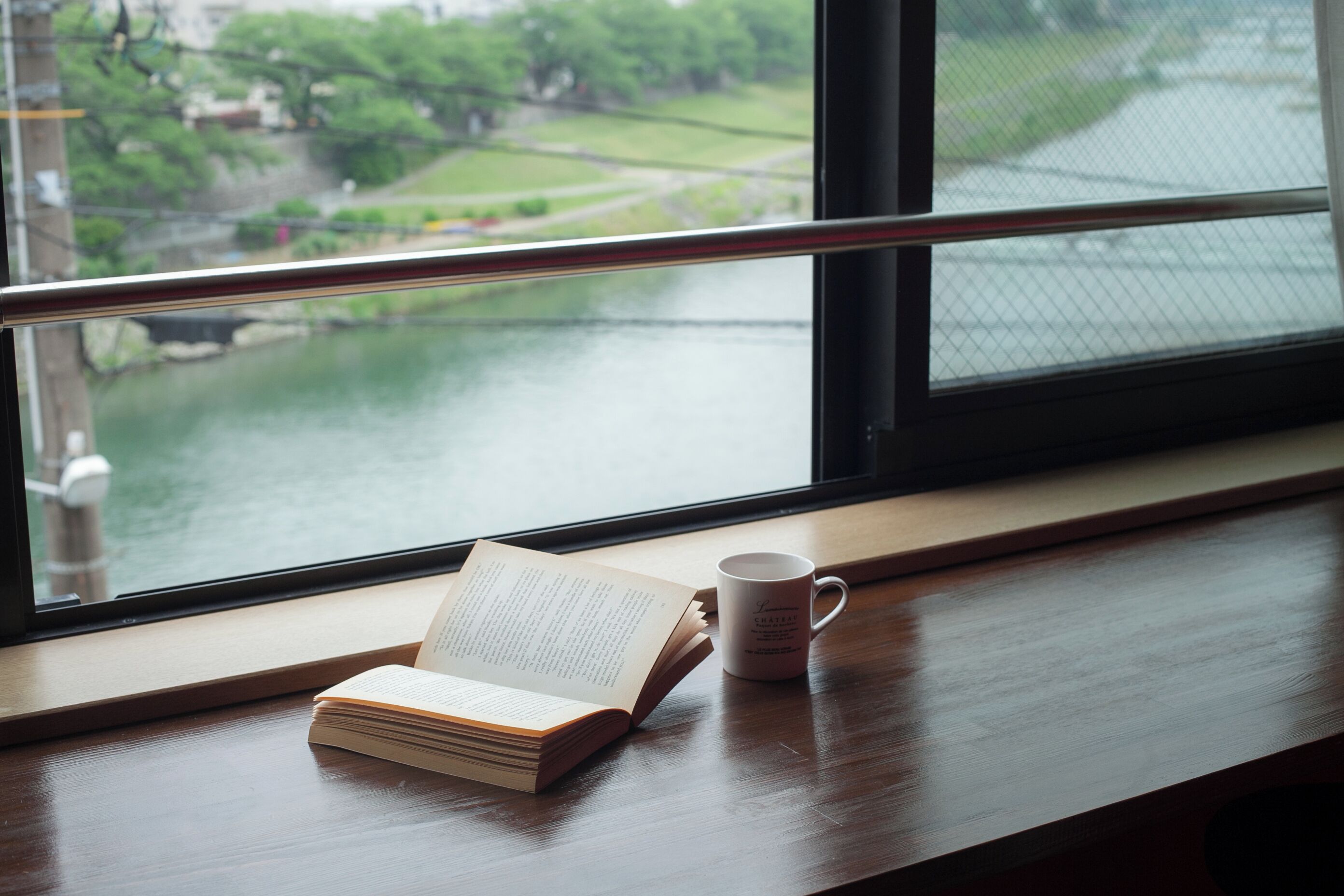 An open book and a mug on a wooden table by a window with a view of greenery and water.