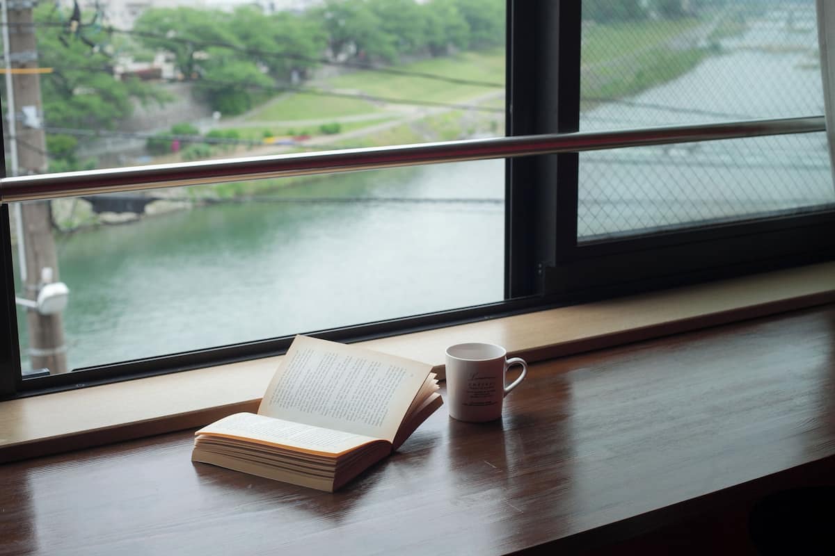 Un livre ouvert et une tasse sur une table en bois près d’une fenêtre donnant sur un paysage verdoyant et une étendue d’eau.