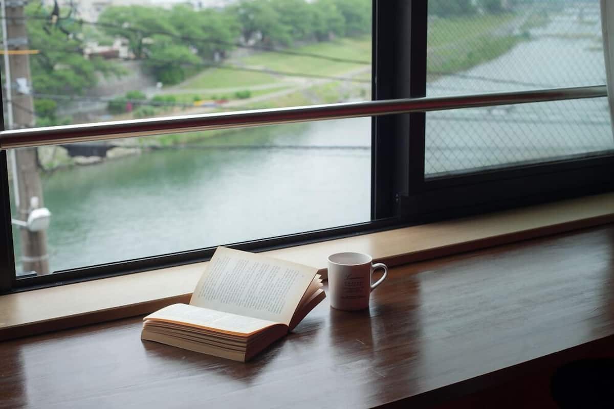 Une fenêtre donnant sur un paysage verdoyant, un livre et une tasse posés sur une étagère en bois.