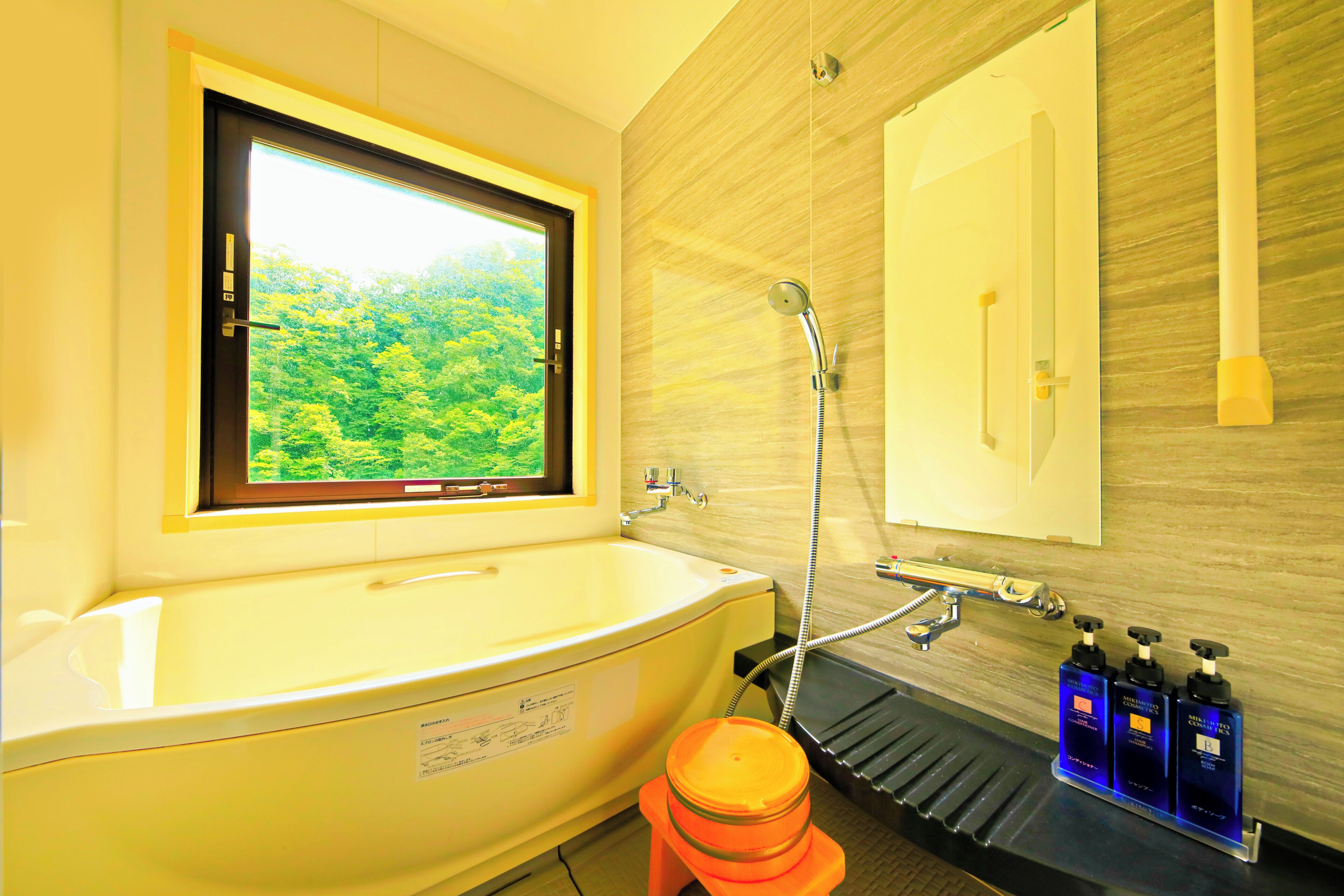 A bathroom with a bathtub, a window with a view of greenery, a shower, and a sink with toiletries.