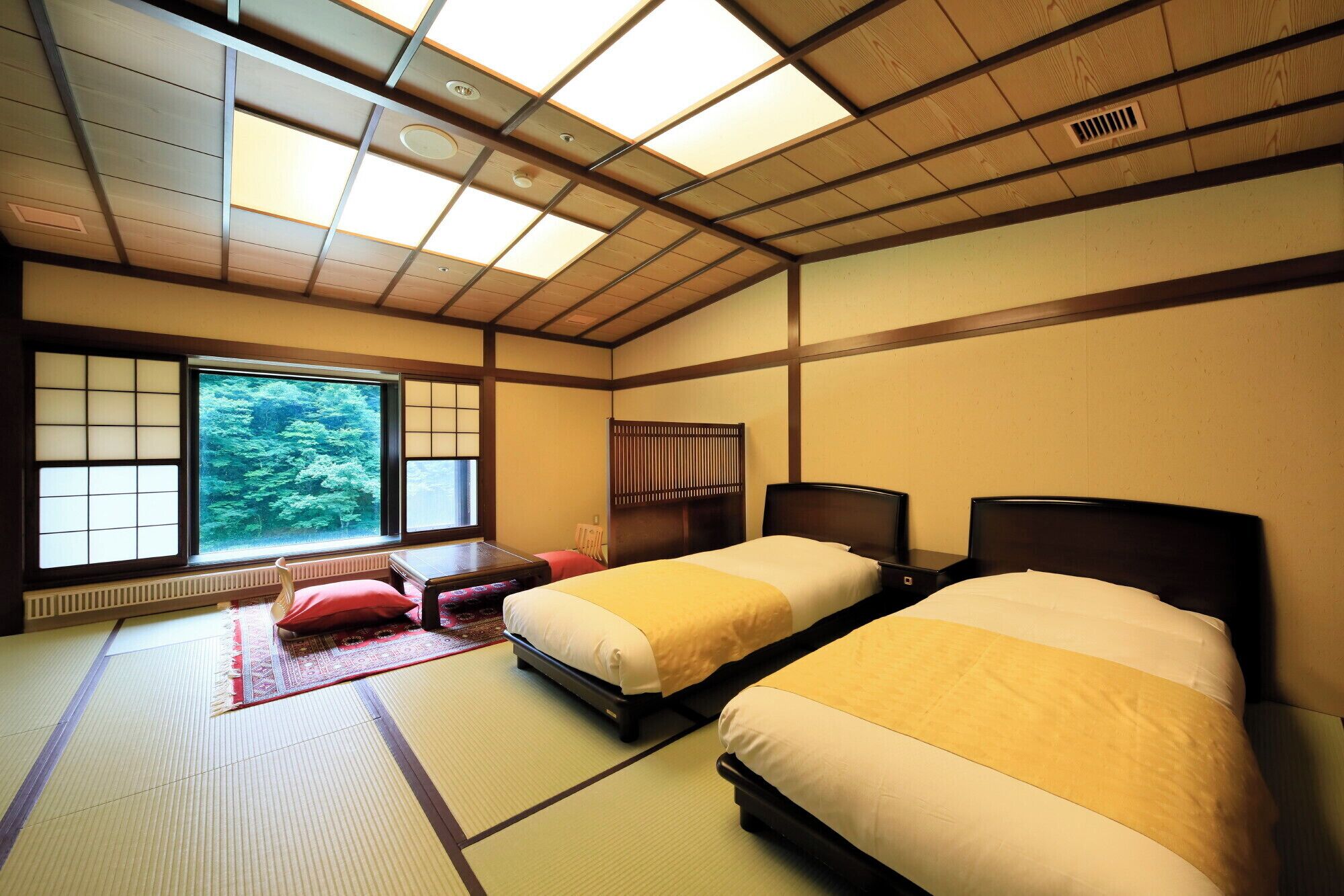A traditional Japanese-style room with tatami flooring, two beds with dark wooden headboards, a large window with shoji screens, and a view of greenery outside.