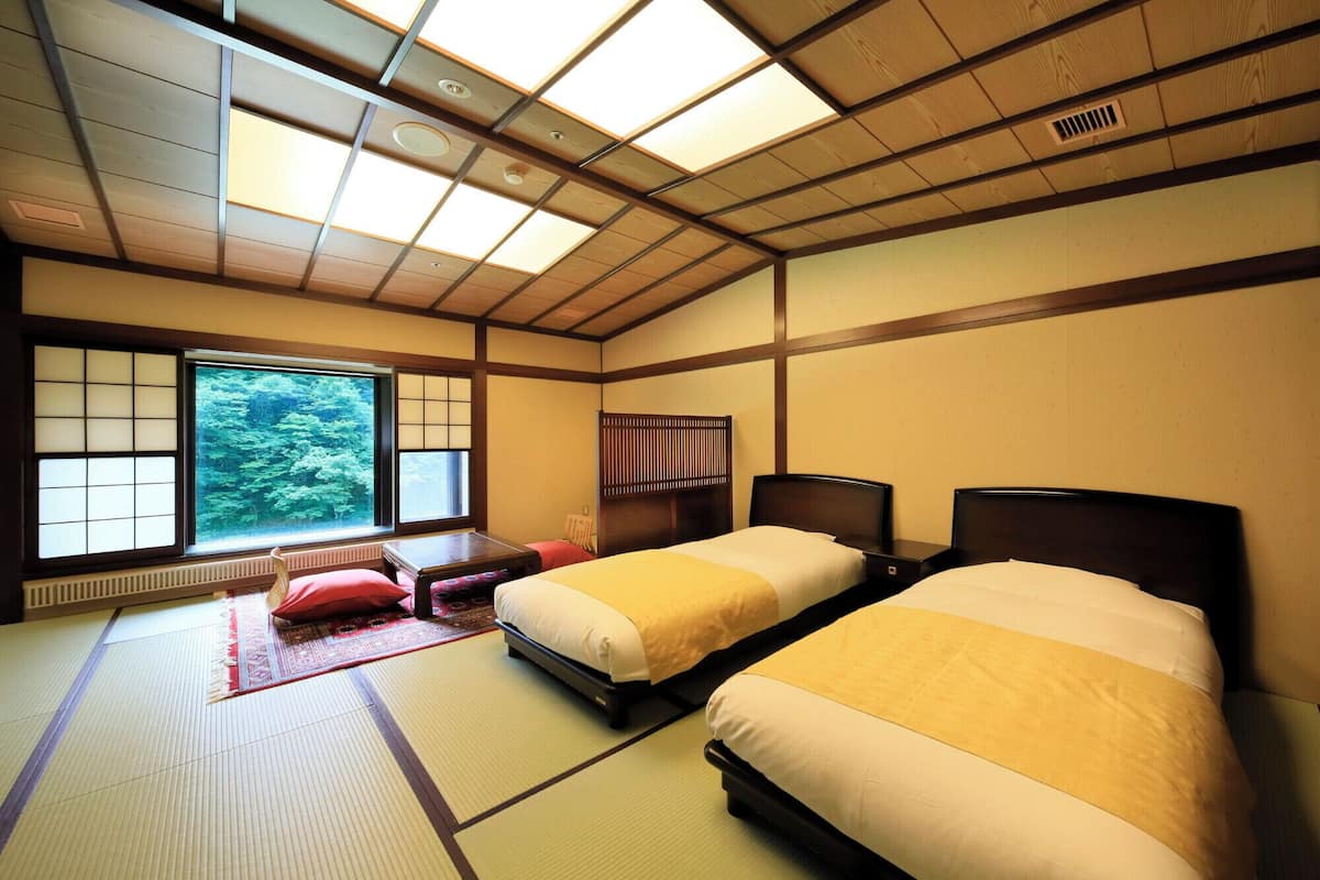 A traditional Japanese-style room with tatami flooring, two beds with dark wooden headboards, a large window with shoji screens, and a view of greenery outside.