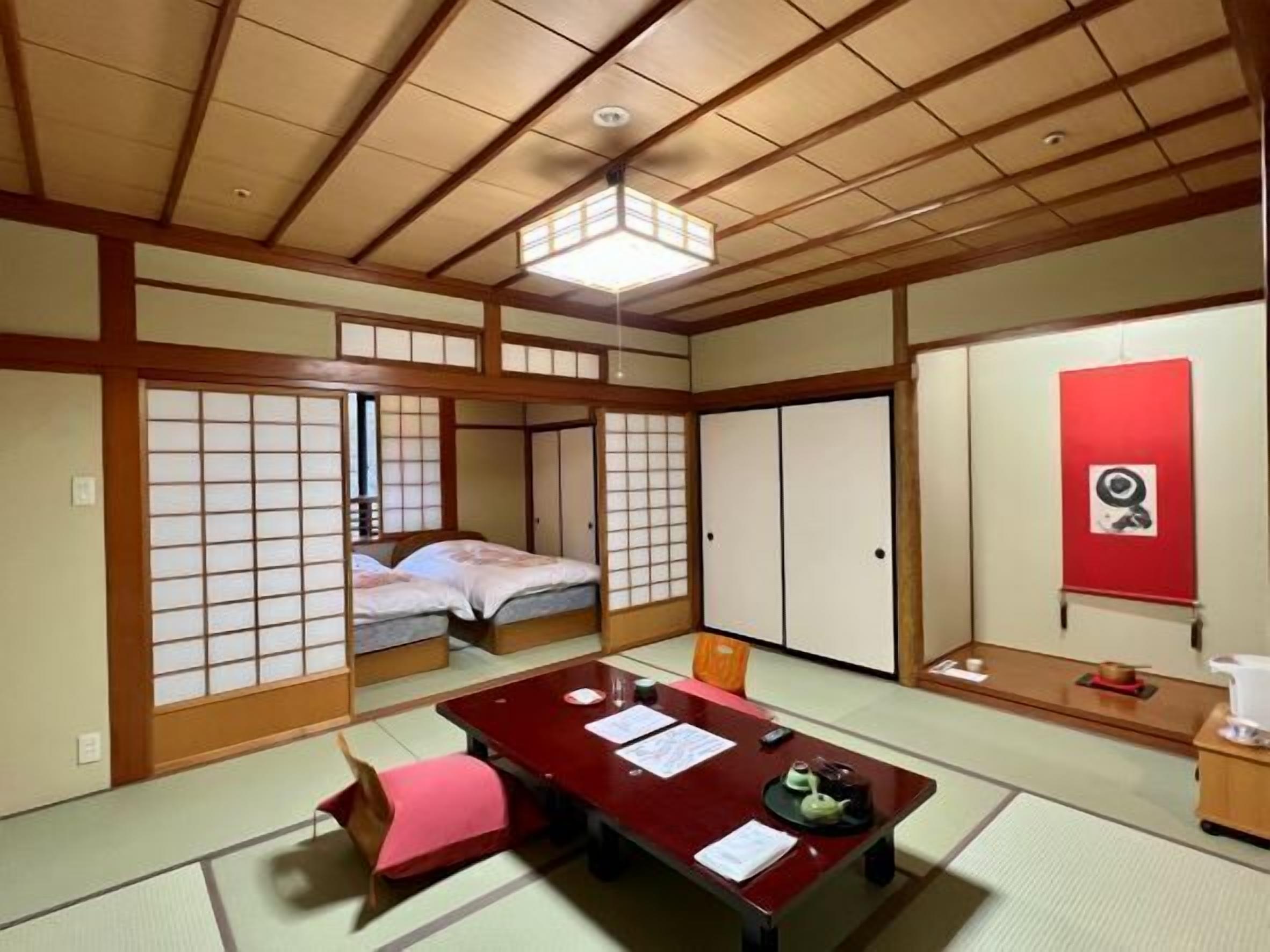 A traditional Japanese room with tatami flooring, a low table with documents, a bed with futon, sliding doors, and a red wall with a picture.