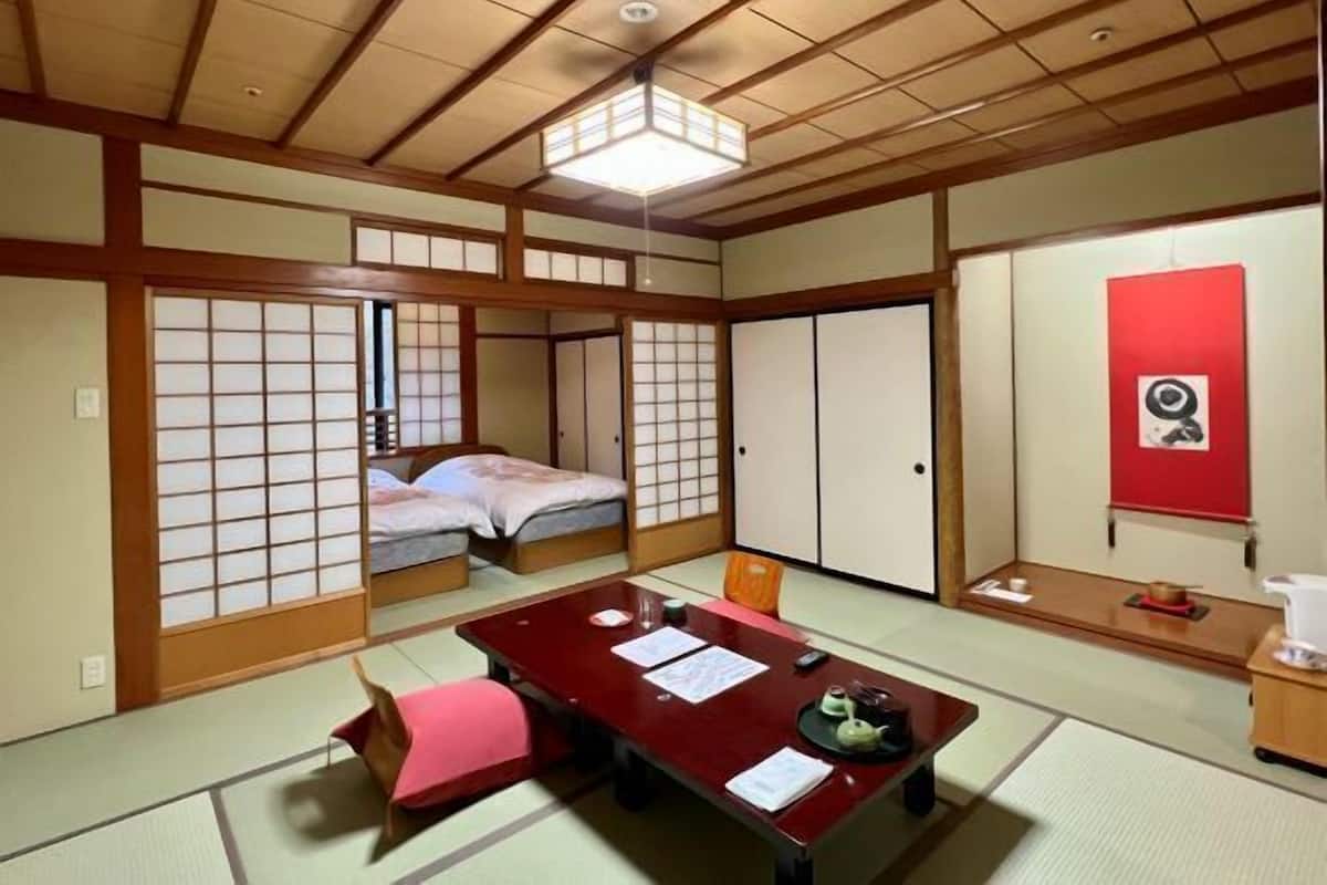 A traditional Japanese room with tatami flooring, a low table with documents, a bed with futon, sliding doors, and a red wall with a picture.