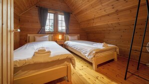 Quarto de cabana aconchegante com duas camas, paredes de madeira, uma janela com cortinas e uma mesa de cabeceira com abajur.