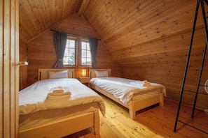 A cozy cabin bedroom with two beds, wooden walls, a window with curtains, and a nightstand with a lamp.