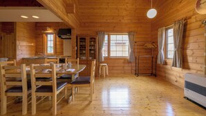 Interior de uma cabana de madeira com mesa de jantar, cadeiras, cozinha e uma janela grande com cortinas.