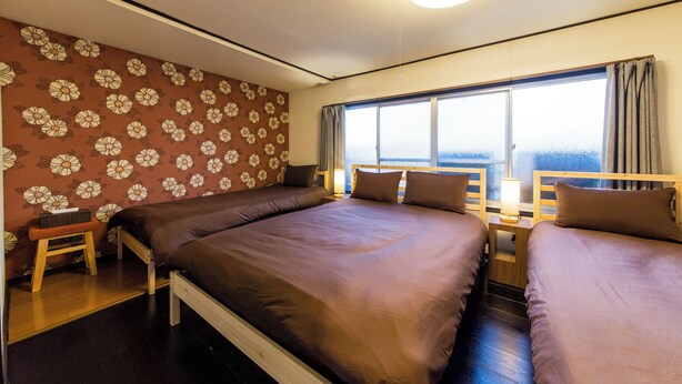 A room with two beds, a wooden nightstand, a small table, and a window with curtains. - Maiko-han Inn (Kyoto)