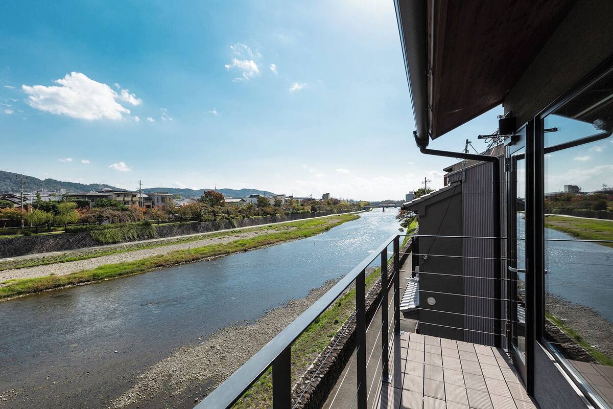 Blick auf einen Fluss von einem Balkon mit einer Steinmauer, einem Holzboden und einem modernen Gebäude mit Metallgeländer.