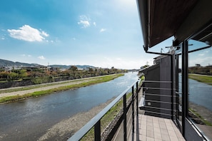 A river view from a balcony with a stone wall, a wooden deck, and a modern building with a metal railing. - Kyomachiya Yaeya Toichicho (Kyoto)