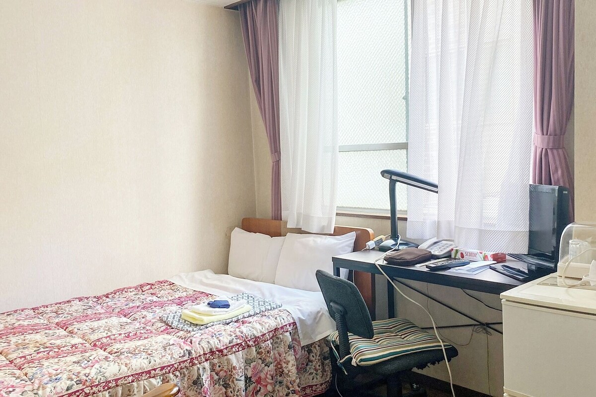 A hotel room with a bed, a desk with a computer, a chair, and a small kitchenette.