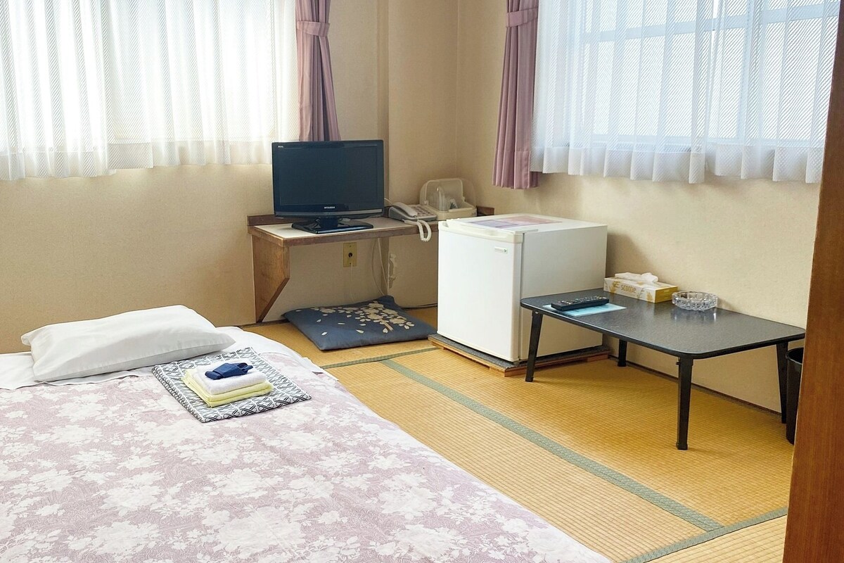 A hotel room with a bed, a desk with a computer, a refrigerator, and a small table with a remote control.
