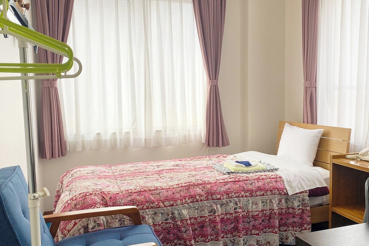 A hotel room with a bed, a desk with a computer, a chair, and a bedside table.