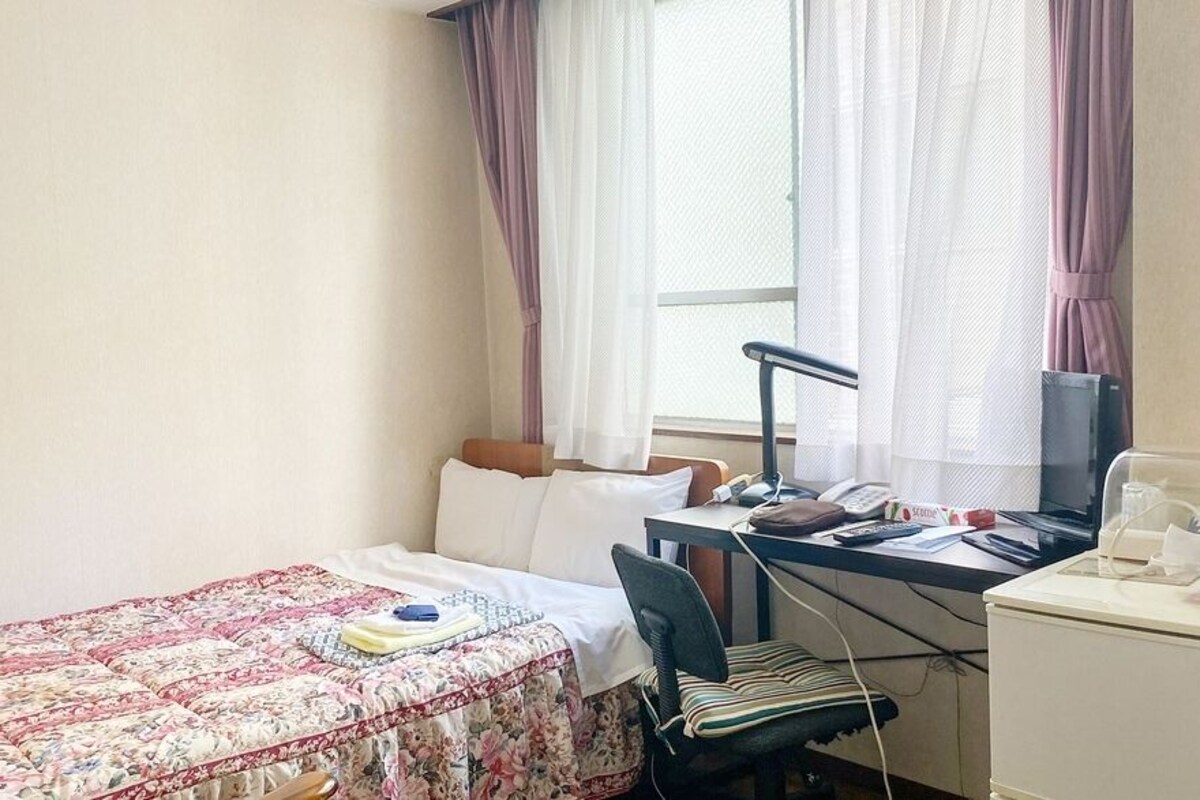 A hotel room with a bed, a desk with a computer, a chair, and a small cabinet.