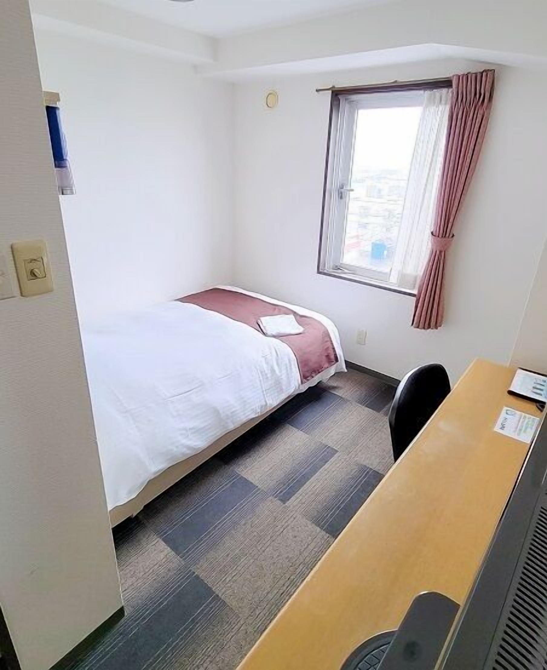 A small hotel room with a single bed, a desk, a chair, and a window with curtains.