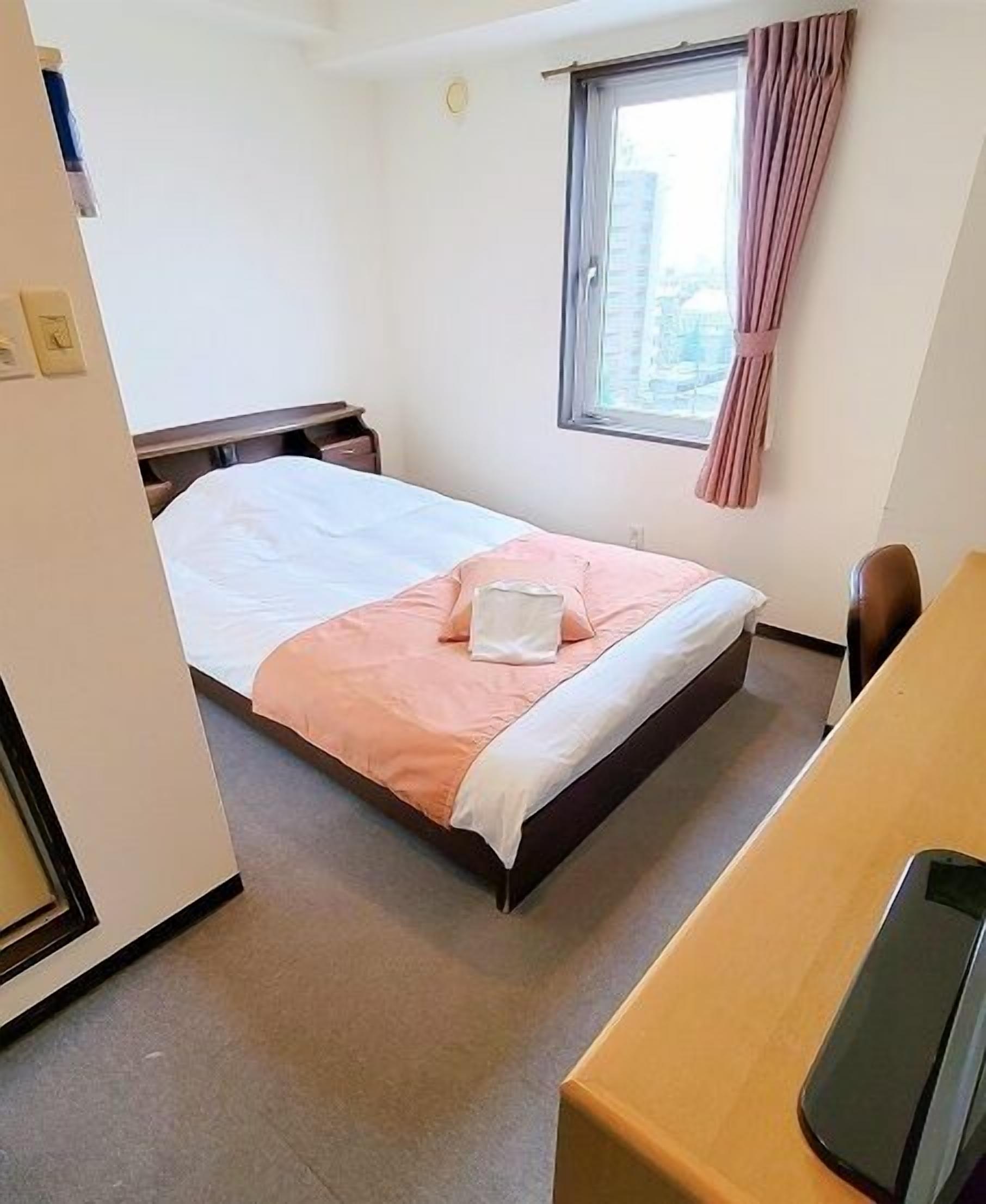 A hotel room with a bed, a desk, a chair, a window with curtains, and a view of buildings outside.
