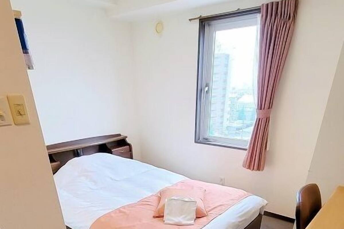 A hotel room with a bed, a desk, a chair, a window with curtains, and a view of buildings outside.