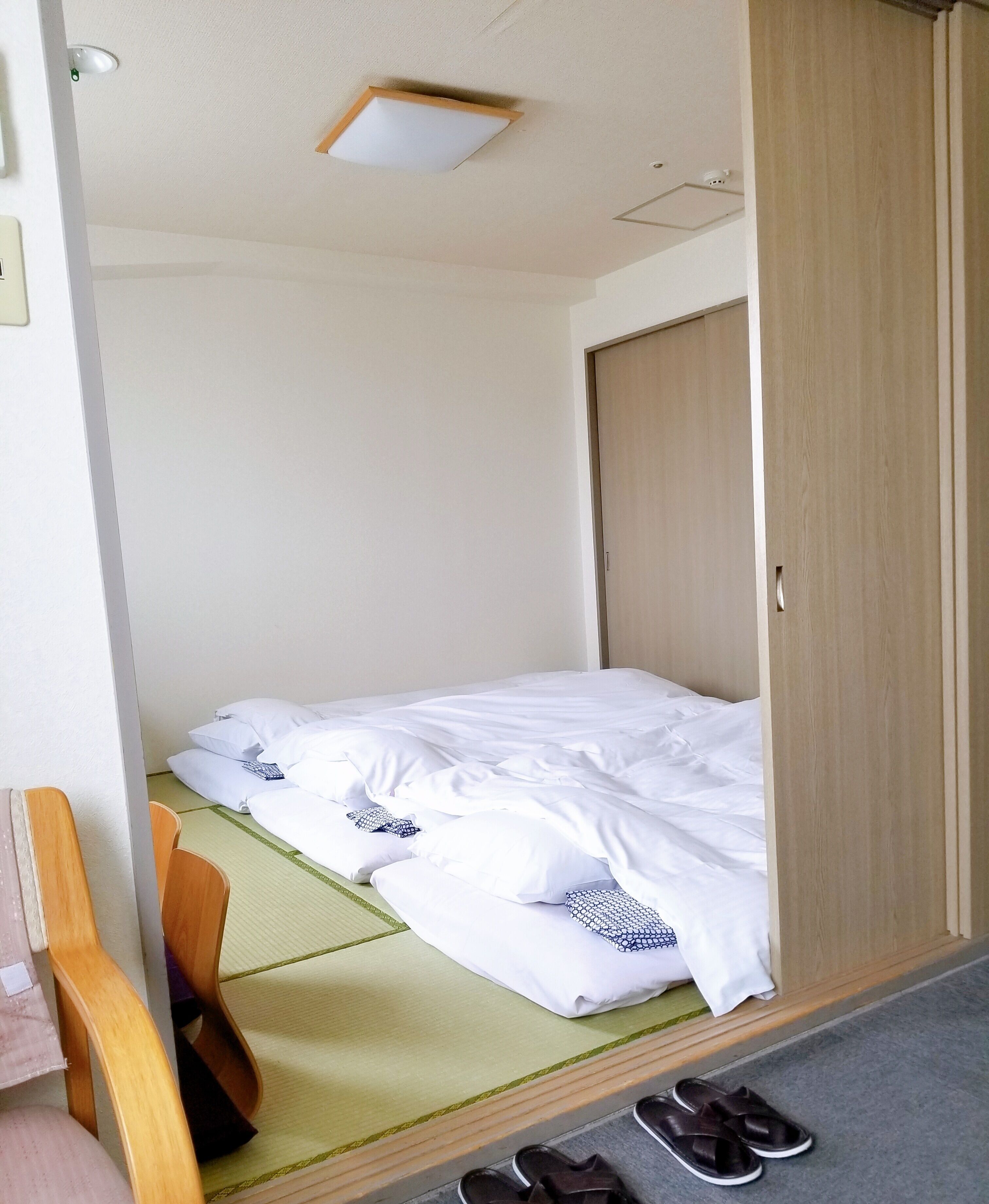 A single bed with white bedding, a wooden wardrobe, and a pair of slippers on the floor.