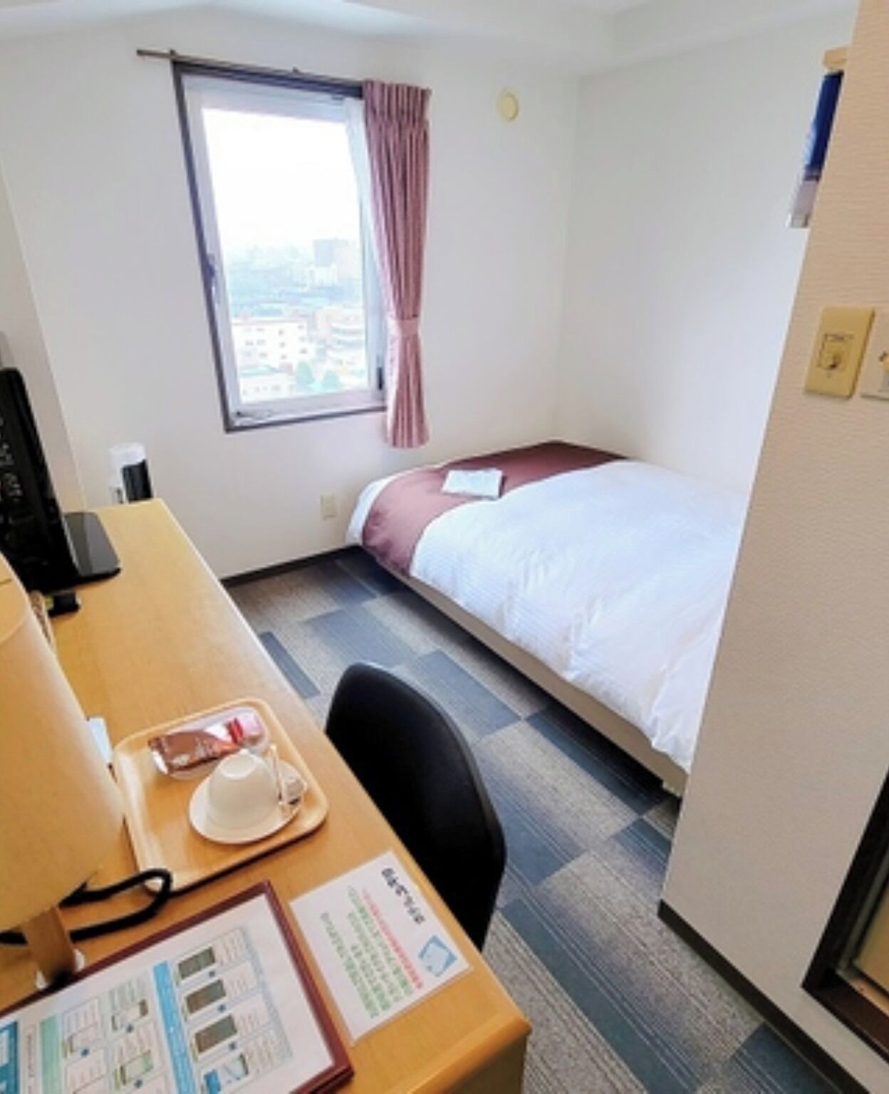 Ein kleines Hotelzimmer mit einem Einzelbett, einem Schreibtisch mit Computer, einem Fenster mit Vorhängen und Blick auf Gebäude.